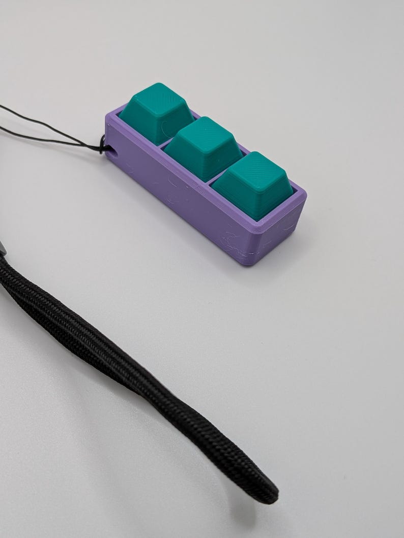 May include: A custom-made, 3D-printed keycap holder with three teal keycaps set in a purple base. A black lanyard is attached for portability. The keycap holder is designed for a specific keyboard or project.