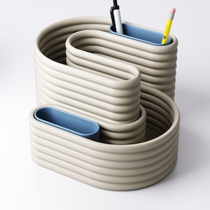 May include: A beige, wave-shaped desk organiser with three blue pen holders. A black pen and a yellow pencil are in the top holder. The organiser is made of a ridged material and sits on a white surface. A modern office supply.