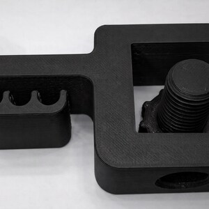 Puede incluir: Una abrazadera negra impresa en 3D con un perno y tres ranuras curvas. La abrazadera tiene forma rectangular con bordes redondeados y un perno roscado para apretar. El objeto est&aacute; dise&ntilde;ado para sujetar o asegurar elementos.