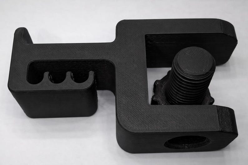 Puede incluir: Una abrazadera negra impresa en 3D con un perno y tres ranuras curvas. La abrazadera tiene forma rectangular con bordes redondeados y un perno roscado para apretar. El objeto est&aacute; dise&ntilde;ado para sujetar o asegurar elementos.