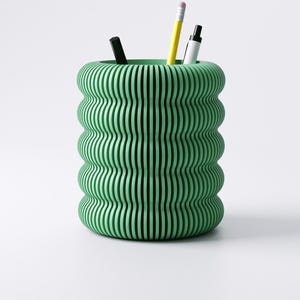 May include: A green desk organizer with a unique, wavy, vertical-line design. The organizer holds a black pen, a yellow pencil, and a white marker. The container is cylindrical and made of a solid material.