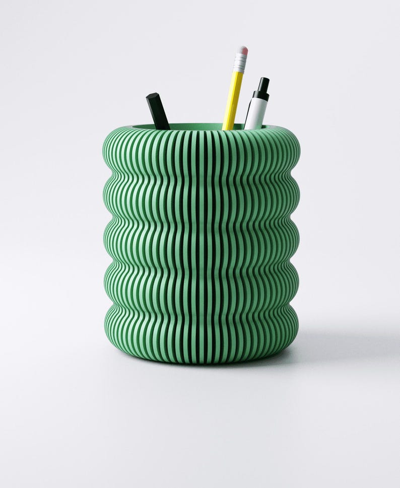 May include: A green desk organizer with a unique, wavy, vertical-line design. The organizer holds a black pen, a yellow pencil, and a white marker. The container is cylindrical and made of a solid material.