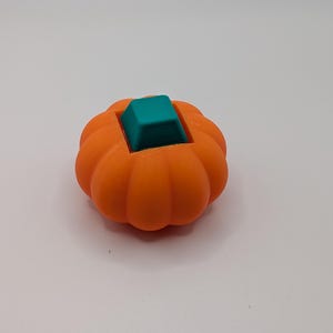 May include: An orange, pumpkin-shaped object with a teal square insert. The pumpkin has a ribbed texture and a rounded shape. The teal insert is a solid, square shape that sits in the center of the pumpkin.