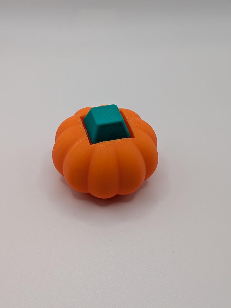 May include: An orange, pumpkin-shaped object with a teal square insert. The pumpkin has a ribbed texture and a rounded shape. The teal insert is a solid, square shape that sits in the center of the pumpkin.
