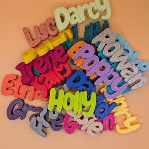 May include: A selection of brightly coloured 3D-printed name cutouts in various fonts. Names such as Darcy, Rowan, and Holly are visible. The cutouts are in shades of green, purple, blue, and pink. The background is a peach colour.