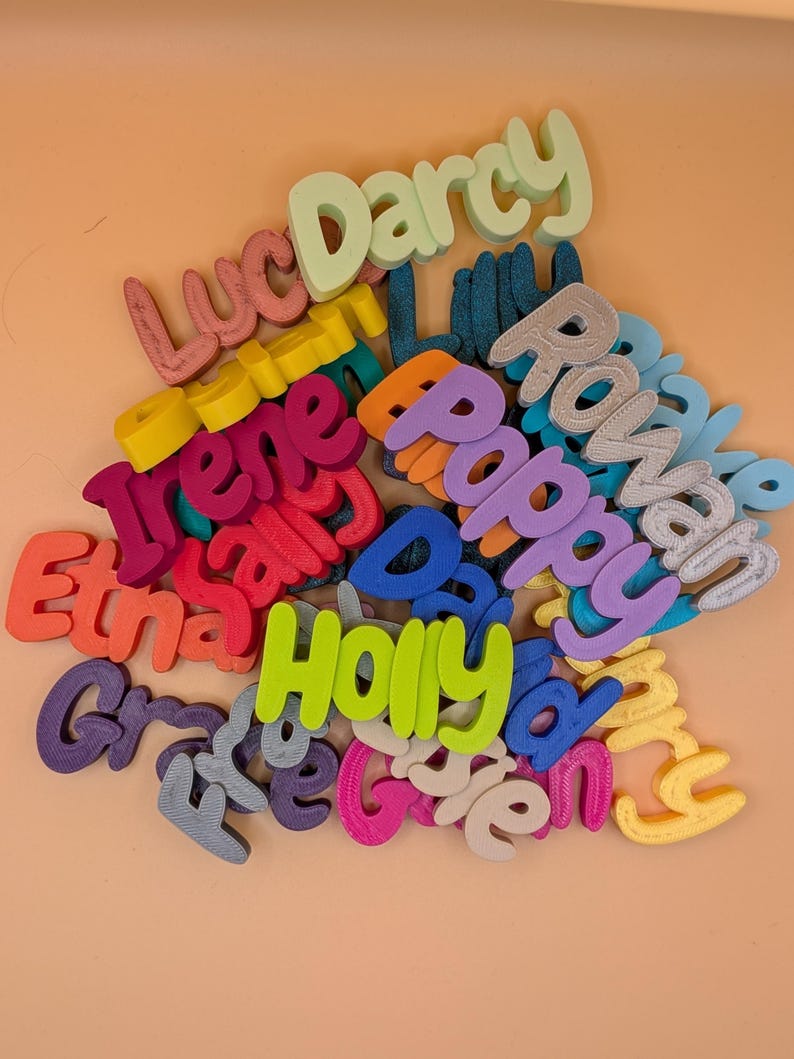 May include: A selection of brightly coloured 3D-printed name cutouts in various fonts. Names such as Darcy, Rowan, and Holly are visible. The cutouts are in shades of green, purple, blue, and pink. The background is a peach colour.