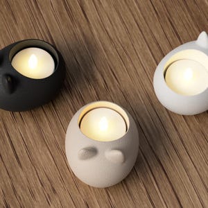 May include: Three ceramic candle holders in black, white, and beige, each with a lit candle inside. The holders have a rounded shape with small, ear-like protrusions. The candles emit a warm, soft glow, creating a cozy ambiance.