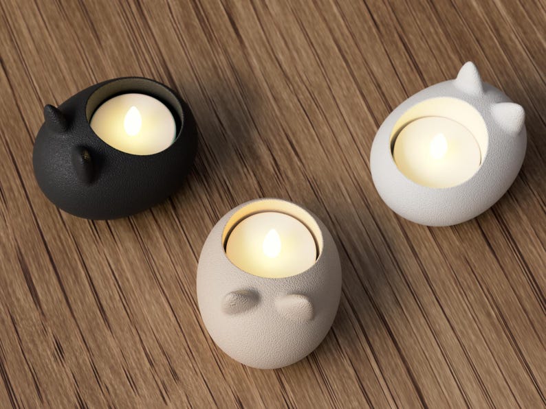 May include: Three ceramic candle holders in black, white, and beige, each with a lit candle inside. The holders have a rounded shape with small, ear-like protrusions. The candles emit a warm, soft glow, creating a cozy ambiance.