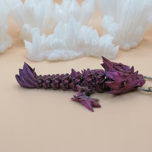 May include: A purple dragon keychain with a silver clasp. The dragon is intricately detailed with a textured surface and a gradient of purple hues. White, crystalline formations are in the background.