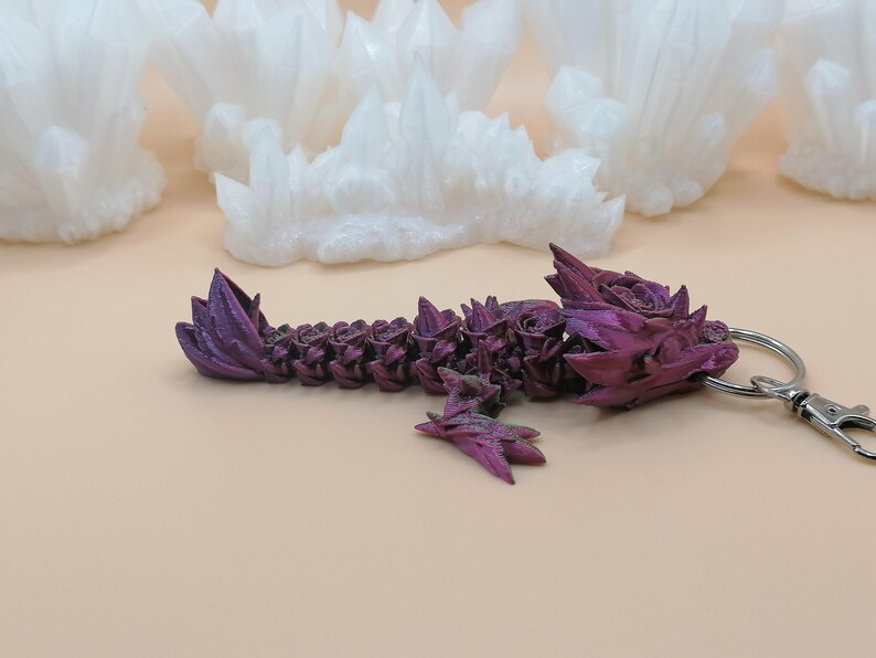 May include: A purple dragon keychain with a silver clasp. The dragon is intricately detailed with a textured surface and a gradient of purple hues. White, crystalline formations are in the background.