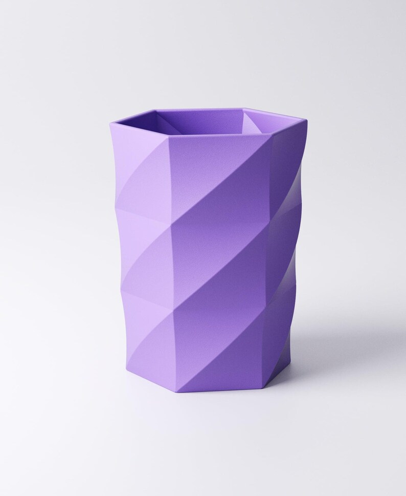 May include: A lavender-colored, geometric pen holder with a faceted design. The hexagonal base transitions into a cylindrical shape with diagonal, textured panels. The interior is divided into sections.