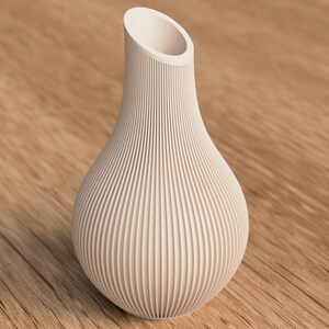 May include: A cream-colored vase with a bulbous base and a narrow, angled opening. The vase has vertical, ridged detailing that covers the entire surface. The vase is made of a solid material and sits on a wooden surface.