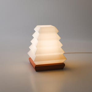 May include: A table lamp featuring a white, pleated, trapezoidal shade that emits a soft, warm glow. The shade is mounted on a solid, square-shaped wooden base. A thin white power cord extends from the side of the base, providing electrical power.