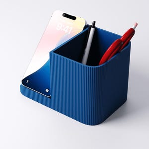May include: A blue desk organizer with a ribbed texture. A smartphone is in the left slot, displaying the time 9:41. A white pen and two red pens are inside the organizer. The organizer is on a white surface.