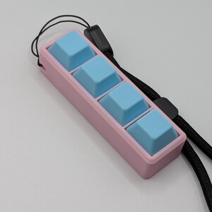 May include: A pink rectangular device with four light blue square buttons. A black lanyard is attached to the device. The device is designed for use with a keyboard.