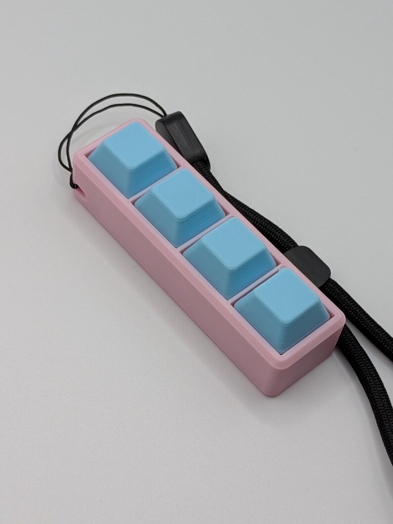 May include: A pink rectangular device with four light blue square buttons. A black lanyard is attached to the device. The device is designed for use with a keyboard.