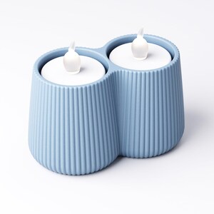 May include: A light blue, double-cup candle holder with two white LED candles. The holder has a ribbed texture and a modern design. The candles have a realistic flame effect, creating a warm and inviting ambiance.