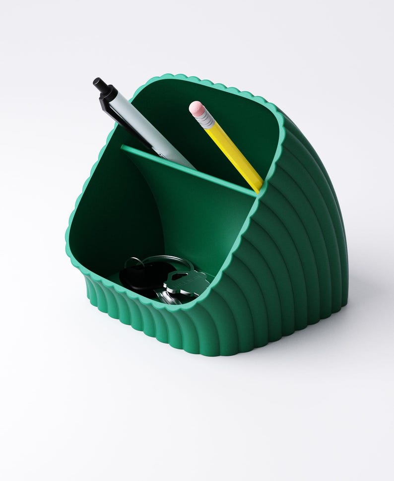 May include: A dark green desk tidy with a scalloped edge design. The organiser has three compartments and contains a black pen, a grey pen, a yellow pencil, and a set of keys. The organiser is made of a solid material.