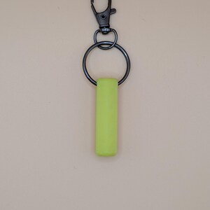May include: A neon green keychain with a black metal clasp and ring. The rectangular green piece is attached to the ring, which connects to the clasp. The background is a solid, neutral beige color.