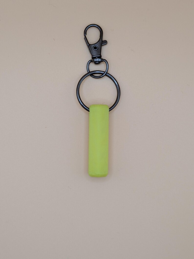 May include: A neon green keychain with a black metal clasp and ring. The rectangular green piece is attached to the ring, which connects to the clasp. The background is a solid, neutral beige color.