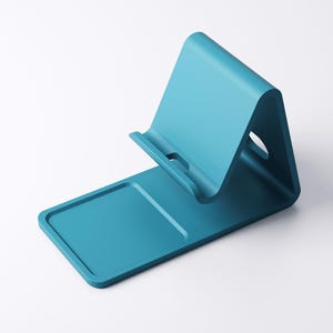 May include: A teal-colored phone stand with a rectangular base and an angled back. The stand has a slot to hold a phone and a small cutout on the back. The design is minimalist and modern.