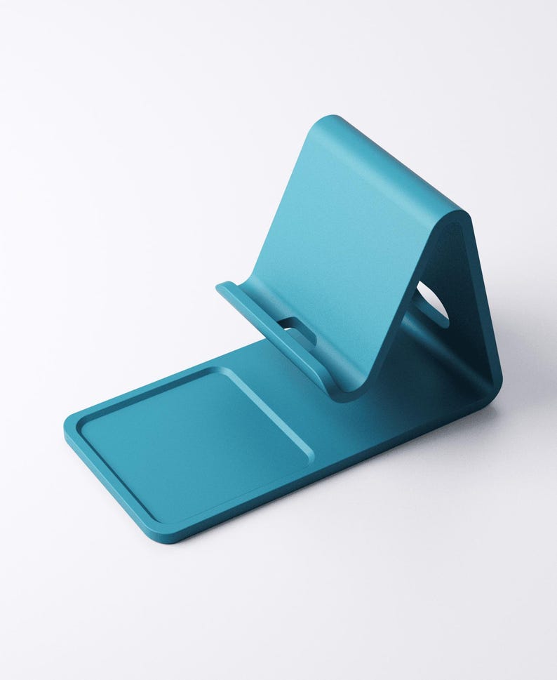 May include: A teal-colored phone stand with a rectangular base and an angled back. The stand has a slot to hold a phone and a small cutout on the back. The design is minimalist and modern.