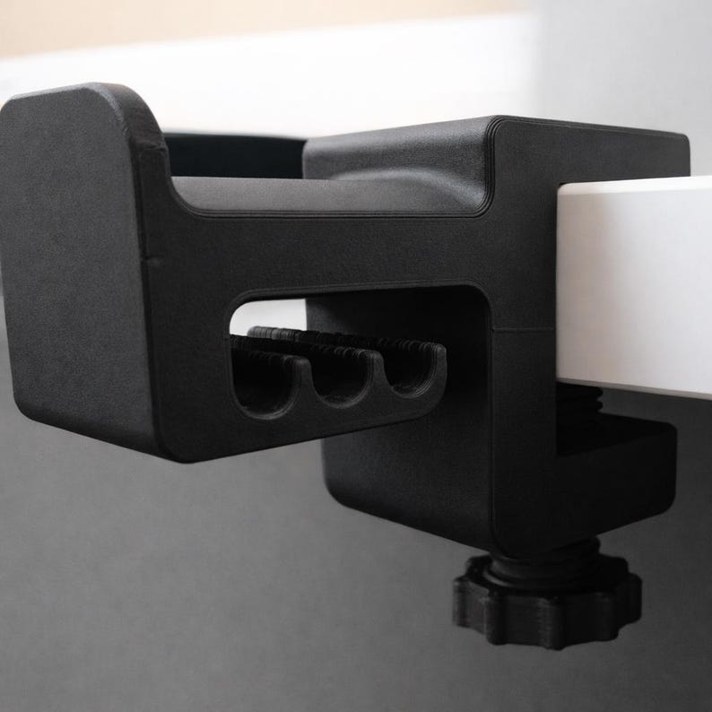 Puede incluir: Un soporte negro para auriculares montado en un escritorio. El soporte tiene una abrazadera para sujetarlo a un escritorio, una parte superior curvada y una secci&oacute;n inferior con tres ranuras curvas. El soporte est&aacute; hecho de un material negro mate.