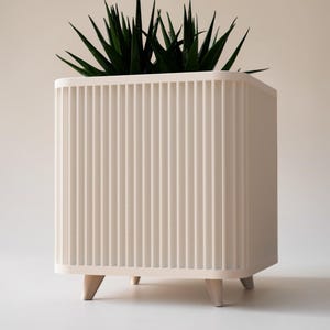 May include: A cream-coloured, square planter with vertical ribbed detailing and short, angled wooden legs. It contains a vibrant green plant with long, pointed leaves. The design is modern and minimalist.