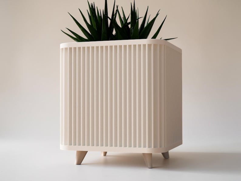 May include: A cream-coloured, square planter with vertical ribbed detailing and short, angled wooden legs. It contains a vibrant green plant with long, pointed leaves. The design is modern and minimalist.