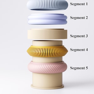 K&ouml;nnte beinhalten: Ein Stapel aus f&uuml;nf farbenfrohen, segmentierten Armb&auml;ndern in Blau-, Lavendel-, Beige-, Gelb- und Rosat&ouml;nen. Jedes Armband hat ein einzigartiges Design mit gerippten, geschwungenen und geometrischen Mustern. Die Segmente sind mit 1 bis 5 beschriftet.
