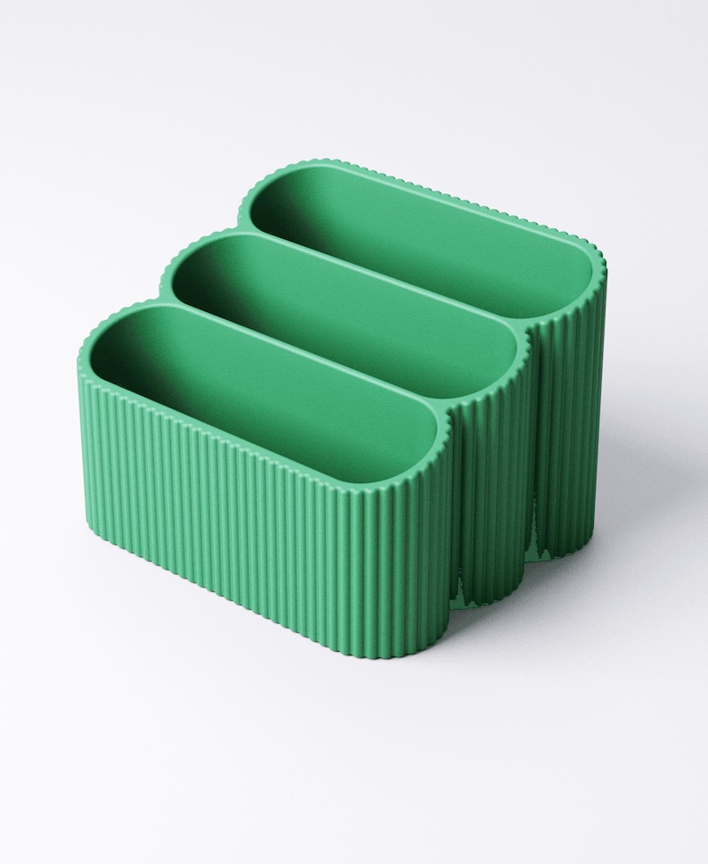 May include: A three-section, green desk organizer with a ribbed exterior. The organizer has an oval shape and is made of a solid, opaque material. The sections are open at the top for easy access.