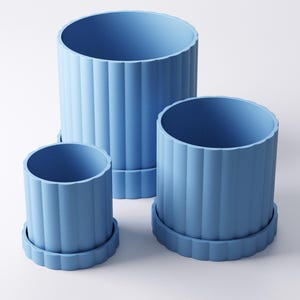 May include: Three light blue cylindrical planters of varying sizes. Each planter has vertical ridged detailing and sits on a matching, scalloped-edge saucer. The planters are arranged on a white surface.