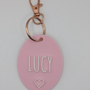 May include: A pink oval keychain with the name "LUCY" in white capital letters and a white heart symbol. The keychain has a gold-colored clasp and ring.