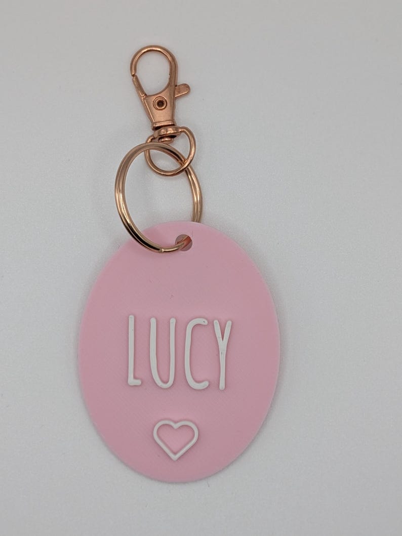 May include: A pink oval keychain with the name "LUCY" in white capital letters and a white heart symbol. The keychain has a gold-colored clasp and ring.