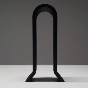 K&ouml;nnte beinhalten: Ein minimalistisches, schwarzmetallenes, bogenf&ouml;rmiges Dekorationsobjekt. Das Objekt hat eine rechteckige Basis mit zwei vertikalen St&uuml;tzen, die sich oben w&ouml;lben und einen Bogen bilden. Der Hintergrund ist ein neutrales Grau.