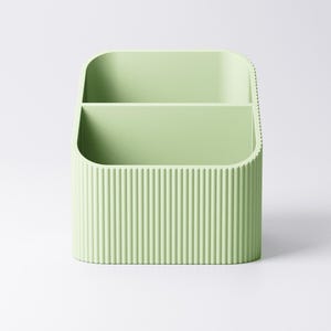 May include: A light green desk organizer with two compartments. The exterior features vertical ribbed detailing. The organizer is made of a solid material and has rounded corners.