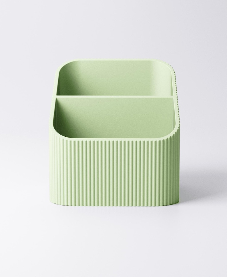 May include: A light green desk organizer with two compartments. The exterior features vertical ribbed detailing. The organizer is made of a solid material and has rounded corners.