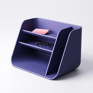 May include: A purple desk organizer with two shelves and a ribbed side. A pink eraser and silver paper clips rest on the shelves. The organizer is made of a solid material and has a modern design.