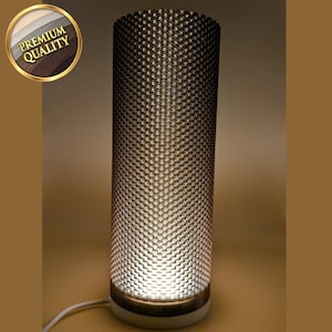 May include: A cylindrical table lamp with a textured, patterned shade. The lamp emits a warm glow, and has a white base. The lamp is plugged in with a white cord. A gold seal with the words "PREMIUM QUALITY" is in the upper left corner.