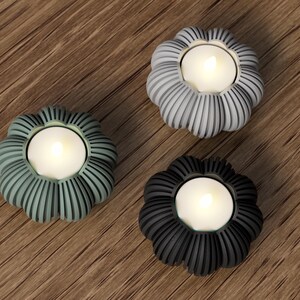 May include: Three decorative candle holders in shades of gray, green, and black. Each holder has a flower-like shape with vertical ribbed detailing and holds a lit, white tea light candle. The holders are arranged on a wooden surface.