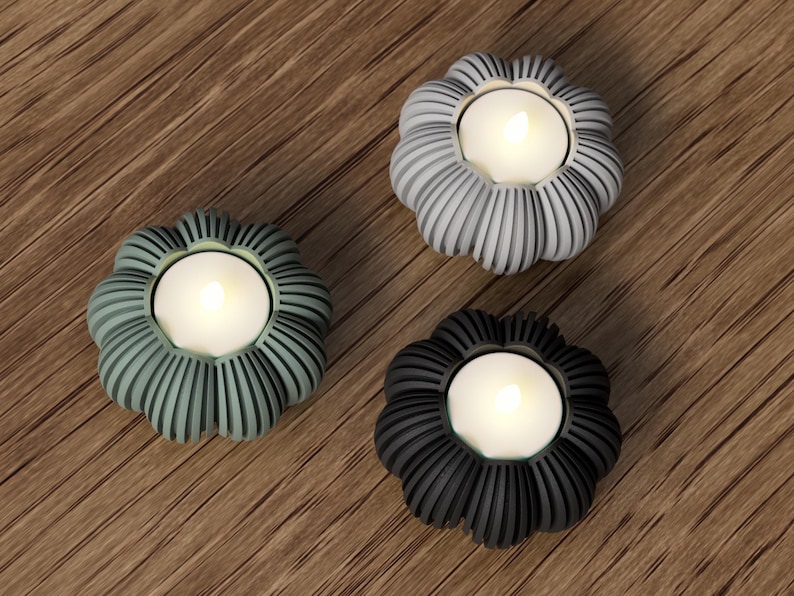 May include: Three decorative candle holders in shades of gray, green, and black. Each holder has a flower-like shape with vertical ribbed detailing and holds a lit, white tea light candle. The holders are arranged on a wooden surface.