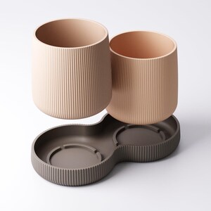 May include: Two beige, ribbed cylindrical cups sit above a dark brown, double-bowl base. The cups have a smooth interior and are designed to fit into the base. The base has a ribbed exterior and a curved shape.