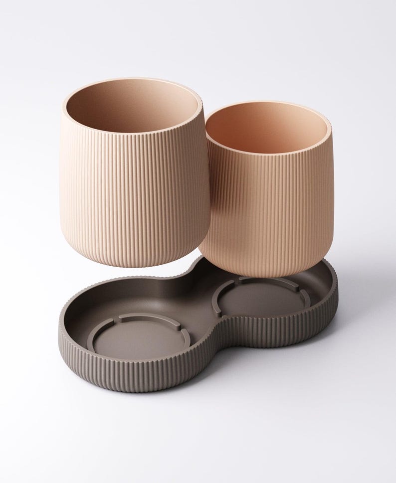 May include: Two beige, ribbed cylindrical cups sit above a dark brown, double-bowl base. The cups have a smooth interior and are designed to fit into the base. The base has a ribbed exterior and a curved shape.