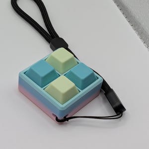 May include: A pastel-colored fidget toy with four square buttons in light blue and pale green. The toy is housed in a square frame with a gradient of pink, blue, and light blue. A black lanyard is attached for carrying.