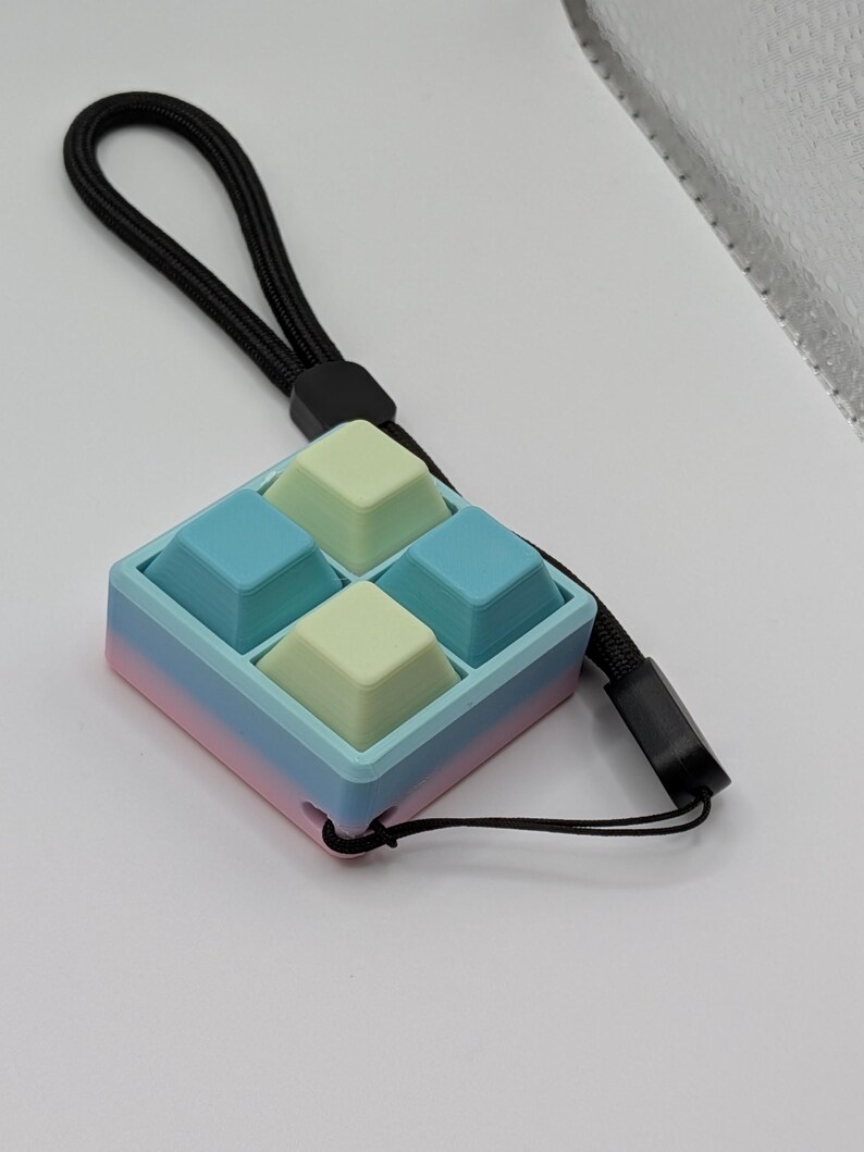 May include: A pastel-colored fidget toy with four square buttons in light blue and pale green. The toy is housed in a square frame with a gradient of pink, blue, and light blue. A black lanyard is attached for carrying.