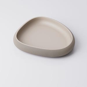 Puede incluir: Una bandeja de servir beige de forma org&aacute;nica. La bandeja tiene un interior liso y poco profundo y un borde texturizado y acanalado. Las ranuras son verticales y crean un patr&oacute;n de rayas alrededor del borde de la bandeja. La bandeja est&aacute; sobre un fondo blanco.