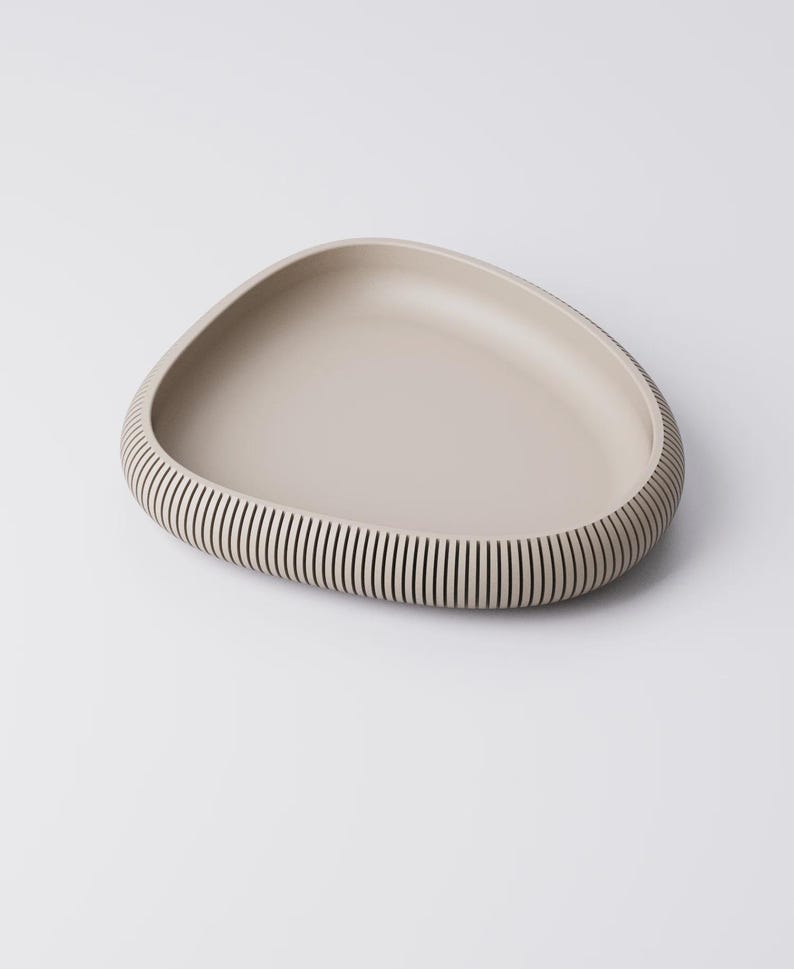 Puede incluir: Una bandeja de servir beige de forma org&aacute;nica. La bandeja tiene un interior liso y poco profundo y un borde texturizado y acanalado. Las ranuras son verticales y crean un patr&oacute;n de rayas alrededor del borde de la bandeja. La bandeja est&aacute; sobre un fondo blanco.