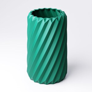 May include: A teal-colored, cylindrical vase with a geometric, ridged design. The vase has a twisted, spiral appearance, with a jagged edge at the top. The vase is made of a solid material and is suitable for home decor.