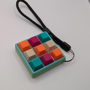 May include: A colorful, square-shaped keyboard keycap set with nine keys in a 3x3 grid. The keys are teal, orange, magenta, and beige. The frame is teal with a rainbow gradient. A black wrist strap is attached.