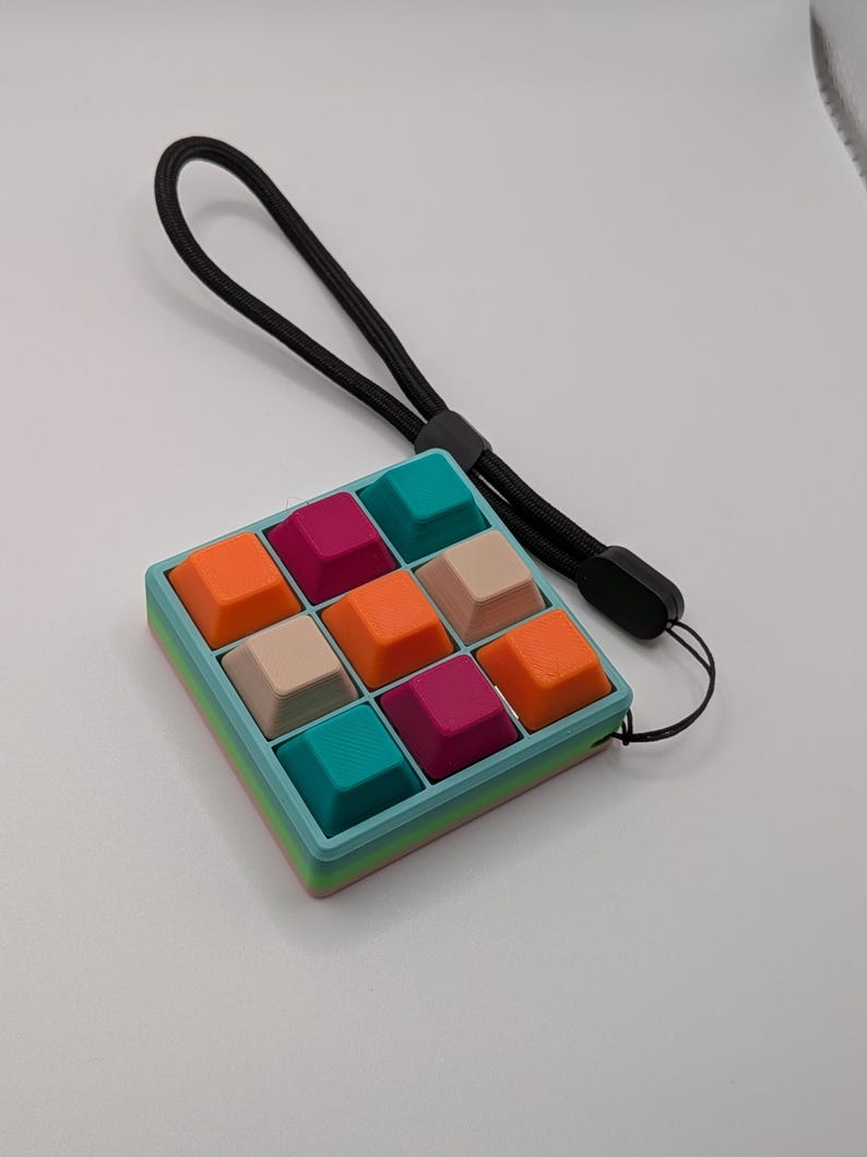 May include: A colorful, square-shaped keyboard keycap set with nine keys in a 3x3 grid. The keys are teal, orange, magenta, and beige. The frame is teal with a rainbow gradient. A black wrist strap is attached.